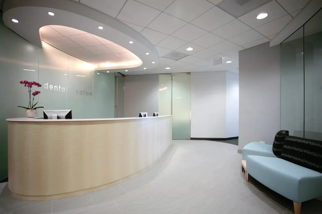 Reception area at Dental Salon Schaumburg: a curved front desk in light wood with a white countertop, the Dental Salon name on a frosted glass wall behind it, and a blue bench in the waiting area.